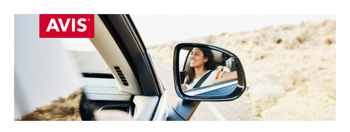 차량 내부에서 바깥 풍경을 바라보는 장면입니다. 사이드미러에는 햇빛이 비치는 사막 도로를 달리며 미소 짓는 여성 운전자의 모습이 비치고 있습니다. 좌측 상단에는 AVIS 로고가 표시되어 있으며, 자유롭고 여유로운 여행 분위기를 전달하는 이미지입니다.